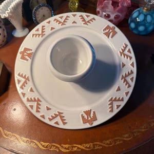May include: White ceramic chip and dip serving dish with a central dip bowl. The rim features a repeating geometric design in a warm brown color. The dish is round and sits on a dark brown surface.