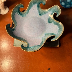 May include: A unique, handcrafted ceramic bowl with a wavy, organic shape. The bowl features a light blue interior with a teal green rim and gold trim. The bowl is a decorative piece, perfect for displaying small items.