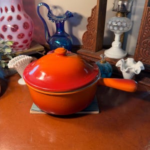 May include: An orange enameled cast iron saucepan with a lid and a single handle. The saucepan is sitting on a small square coaster. Other vintage items are in the background, including vases and a lamp.