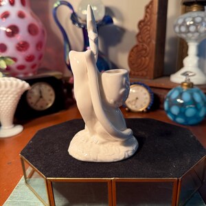 May include: A white ceramic figurine of a hand holding a pipe. The hand is stylized with a long, pointed index finger. The figurine has a smooth, glossy finish and sits on a black velvet surface.
