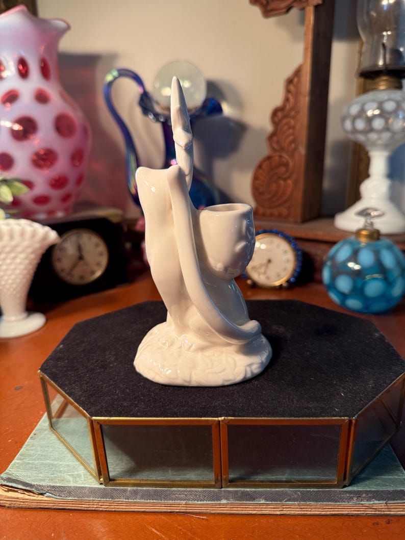 May include: A white ceramic figurine of a hand holding a pipe. The hand is stylized with a long, pointed index finger. The figurine has a smooth, glossy finish and sits on a black velvet surface.