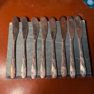 May include: A set of eight vintage butter knives with floral detailing on the handles. The knives are arranged on a small, rectangular, green book. The blades are silver-toned, and the handles have an ornate, embossed design.