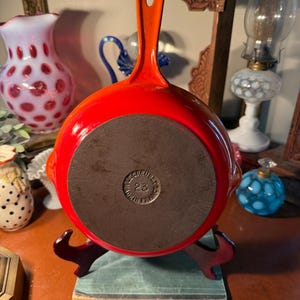 May include: A vibrant red enameled cast iron skillet with an orange handle. The skillet is round with a dark brown cooking surface and the text "Le Creuset 23 Made in France" is visible on the bottom. The skillet is displayed on a decorative stand.