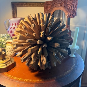 May include: A decorative sphere made of natural driftwood pieces. The sphere is brown and has a spiky, textured appearance. It sits on a round, dark brown table with a decorative border. The background includes a framed mirror and other decorative items.