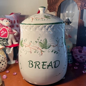May include: White ceramic bread storage container with a green and red floral design and the word "BREAD" written in green. The container has a lid and a scalloped edge.