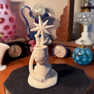 May include: A white ceramic candle holder featuring a star design. The candle holder has a circular base and a curved handle. The star is at the top of the candle holder. The candle holder is on a black surface.