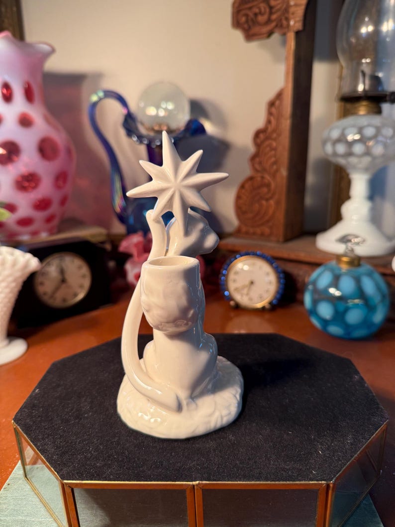 May include: A white ceramic candle holder featuring a star design. The candle holder has a circular base and a curved handle. The star is at the top of the candle holder. The candle holder is on a black surface.