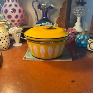 May include: A yellow enamel pot with a lid and silver handles. The pot features a white leaf pattern around the sides. The pot is sitting on a small, green and blue square. The pot is part of a vintage kitchenware collection.