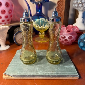 May include: Two vintage yellow glass salt and pepper shakers with silver-colored tops. The shakers have a crackle glass design and sit on a small, aged book. Other antique glass pieces are in the background.