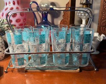 May include: A vintage set of eight clear drinking glasses with a teal and white starburst design, displayed in a chrome metal carrier. The glasses are arranged on a brown surface, suggesting a retro aesthetic.
