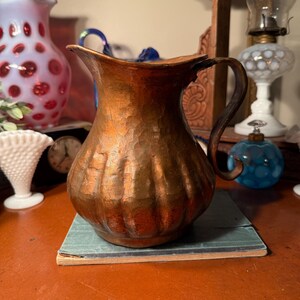 May include: A hammered copper pitcher with a curved handle and a wide spout. The pitcher has a textured surface and sits on a small, rectangular book. Other antique items are in the background, including a pink and white vase.