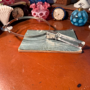 May include: A vintage silver-toned metal magnifying glass with a clear, rectangular handle. The magnifying glass is resting on a small, aged, green book. The background features antique glass and clocks.