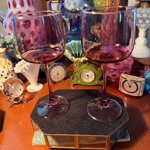 May include: Two matching wine glasses crafted from translucent purple glass. Each glass features a smooth, rounded bowl, a slender stem, and a circular base. The glasses are positioned upright, showcasing the consistent deep violet hue of the glass material.