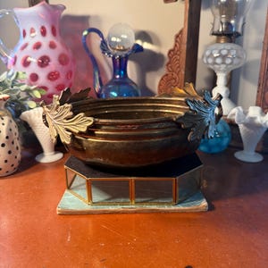 May include: A hammered brass bowl with leaf accents, resting on a geometric display case. The bowl is a warm gold color, and the leaves are silver. Other vintage glass pieces are in the background.