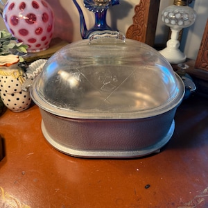 May include: A vintage silver-toned casserole dish with a clear glass lid. The dish has a rounded shape and a textured exterior. The lid features a handle and a diamond pattern. The dish is likely made of metal and glass.
