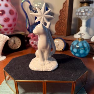May include: A white ceramic cat figurine holding a star. The cat is seated on a base with leaf detailing. The figurine is displayed on a black velvet-covered, gold-framed display case. Other vintage items are in the background.
