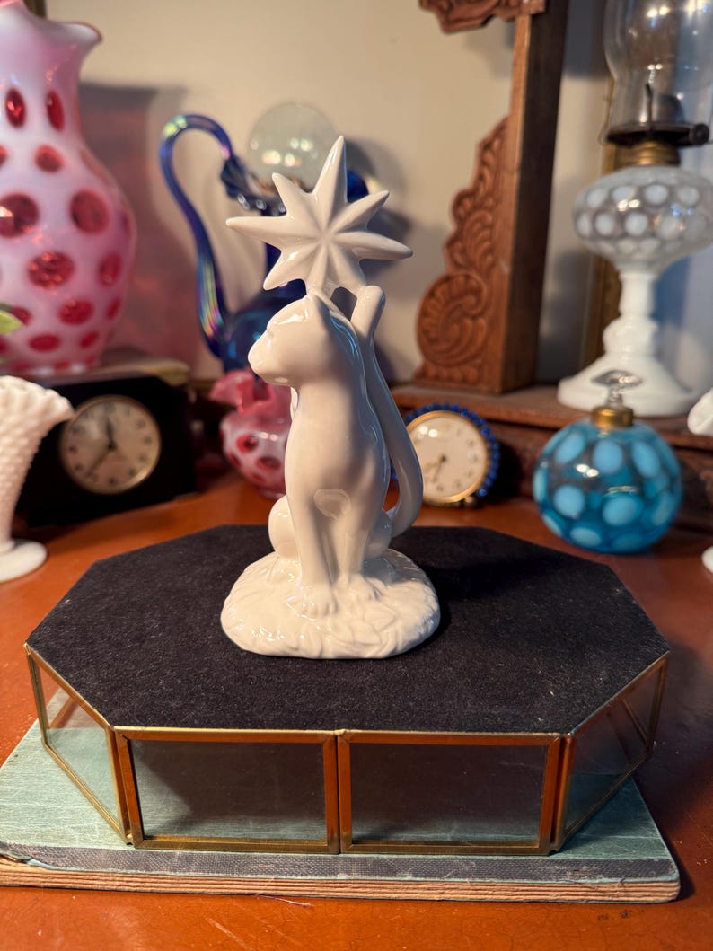 May include: A white ceramic cat figurine holding a star. The cat is seated on a base with leaf detailing. The figurine is displayed on a black velvet-covered, gold-framed display case. Other vintage items are in the background.