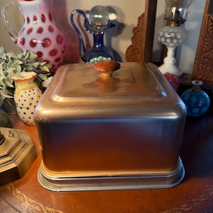 May include: A copper-toned metal bread box with a rounded rectangular shape and a wooden knob on top. The box sits on a silver-colored base. The bread box is displayed on a brown table with other decorative items in the background.