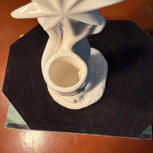 May include: A white ceramic candle holder with a starburst design. The candle holder has a round base and a central opening for a candle. The starburst is at the top of the holder. The candle holder is on a black surface.