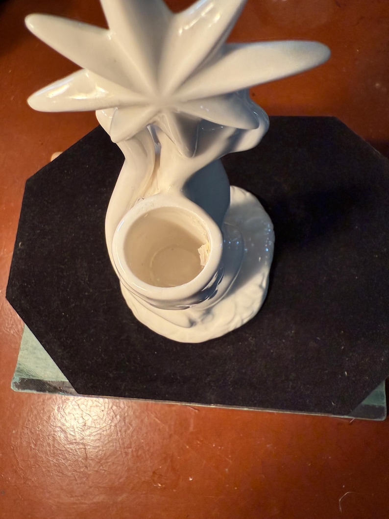 May include: A white ceramic candle holder with a starburst design. The candle holder has a round base and a central opening for a candle. The starburst is at the top of the holder. The candle holder is on a black surface.