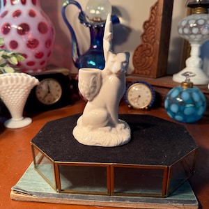 May include: A white ceramic cat figurine with a raised paw, holding a small container. The cat is perched on a black, octagonal display case with gold trim. Other antique items are in the background, including clocks and vases.