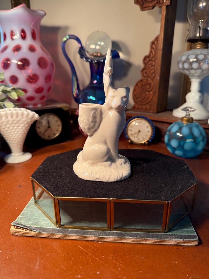May include: A white ceramic cat figurine with a raised paw, holding a small container. The cat is perched on a black, octagonal display case with gold trim. Other antique items are in the background, including clocks and vases.