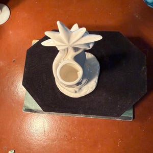 May include: A white ceramic candle holder with a star-shaped design on top. The candle holder has a round base and a cylindrical opening for a candle. The piece is placed on a black surface.