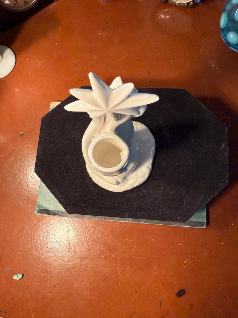 May include: A white ceramic candle holder with a star-shaped design on top. The candle holder has a round base and a cylindrical opening for a candle. The piece is placed on a black surface.