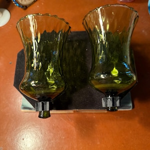 May include: Two green glass candle holders with a ruffled rim. The candle holders are attached to a black base. The base is mounted on a wooden surface. The candle holders have a bulbous base and a flared top.