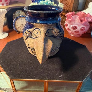 May include: A handmade ceramic vase with a face design. The vase features a dark blue rim and a tan face with blue eyes and a long nose. The vase is sitting on a black surface, with other decorative items in the background.