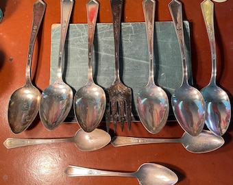 May include: A collection of vintage silverware, including spoons and a fork, arranged on a brown surface. The spoons have ornate handles and rounded bowls. The fork has four tines. The silverware is likely made of silver or a silver-colored metal.