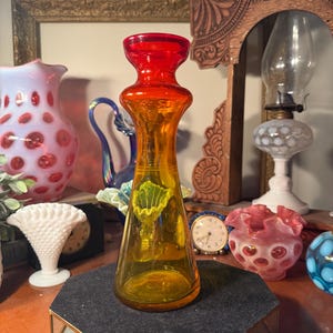 May include: A vibrant, orange and yellow glass vase with a flared base and a rounded top. The vase has a unique shape and is displayed on a black, geometric stand. Other decorative glass pieces are in the background.