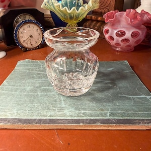 May include: A clear, cut-glass vase with a wide, flared top and a rounded base. The vase sits on a green and gray surface, with other decorative glass pieces and a small clock in the background. The vase has a geometric pattern.