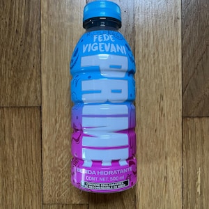May include: A blue and pink plastic bottle of PRIME hydration drink with the text "FEDE VIGEVANI" and "PRIME" on the label. The bottle contains 500 ml and is labeled as a hydrating beverage with sweeteners. It is not recommended for children.