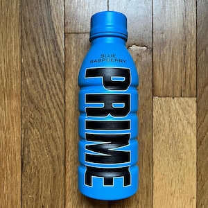 May include: A blue Prime Blue Raspberry drink bottle. The bottle features a bold black and white "PRIME" logo. The top of the bottle is blue, with "BLUE RASPBERRY" printed on the front. The bottle is on a wooden surface.