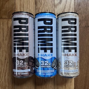 Può includere: Tre lattine di PRIME SHAKE ai gusti cioccolato, biscotti e panna e vaniglia. Ogni lattina è cilindrica, con la parola "PRIME" in grandi lettere nere in grassetto. Le lattine contengono 32g di proteine e sono da 325ml.