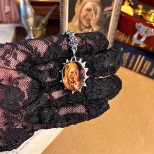 May include: A silver pendant necklace with an oval image of a person in a jester's hat. The pendant has a sunburst design. The necklace is held by a black lace glove.