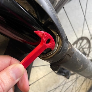 Op de afbeelding: Een rode fietssleutel voor het verwijderen van vorkafdichtingen wordt in een mountainbike-veringvork gestoken. De sleutel wordt door een hand vastgehouden en wordt gebruikt om de vorkafdichtingen te verwijderen. De fiets heeft een zwarte vork.