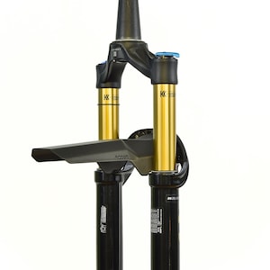 May include: A black and gold mountain bike suspension fork featuring Kashima coated stanchions, a black crown, and a black mudguard attached to the bridge. The fork includes blue adjustment knobs on top and technical labels on the lower legs.