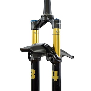 May include: A black and gold mountain bike suspension fork featuring a black mudguard attached to the lower legs. The upper stanchions are gold with Kashima coating, and the lower legs display the number 34 in large gold font with SL text.