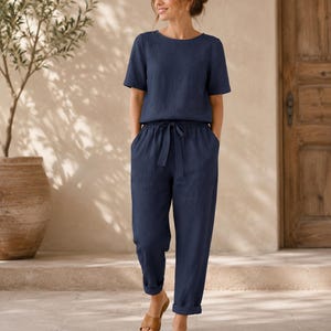 May include: A navy blue two-piece set consisting of a short-sleeved crew neck top and matching trousers. The trousers feature an elasticated waistband with a drawstring tie, side pockets, and cuffed hems. The fabric has a visible textured weave.