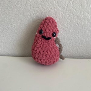 Puede incluir: Un juguete hecho a mano, de felpa, con forma de frijol, en color rosa polvoriento. El juguete tiene ojos negros de botón y una sonrisa negra cosida. Un pequeño trozo de hilo gris se extiende desde el lateral. El juguete está sobre una superficie blanca.