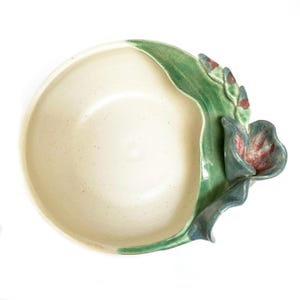 May include: A white ceramic bowl with a green and blue floral design. The bowl has a unique shape with a raised rim and a flower-shaped handle.