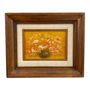 May include: A framed floral painting featuring a bouquet of white and orange flowers in a dark green vase against a yellow background. The painting is signed "Bobby" and is set within a wooden frame.