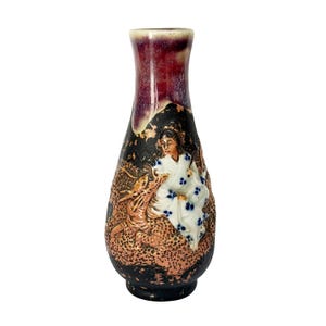 May include: A decorative ceramic vase with a dark red and black glazed finish. The vase features a relief design of a woman in a kimono with blue floral accents and a dragon. The vase is approximately 8 inches tall.