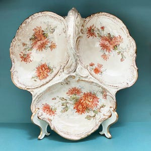 Puede incluir: Fuente de porcelana blanca de tres secciones con asa. La fuente presenta un diseño floral con flores naranjas y verdes, acentuado con ribetes dorados. La fuente está sostenida por un soporte azul claro con detalles dorados.