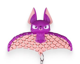 May include: A purple and pink bat-shaped keychain. The bat has large ears, a stern expression, and a segmented wing design. The keychain includes a silver chain and a keyring. The bat is a cartoon-like design.