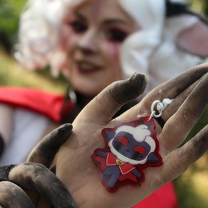 May include: A person wearing a white wig and red clothing is holding a red and white keychain with a cartoon sheep wearing a red cape. The sheep has black eyes and a black mouth.