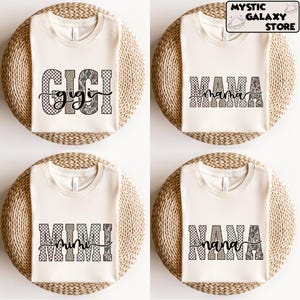 May include: Four cream-coloured t-shirts with different text designs. The shirts feature the words "GIGI", "MAMA", "MIMI", and "NANA" in a mix of patterned fonts, including checkered, leopard print, and script. The shirts are displayed on a woven surface.