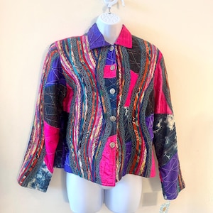 Op de afbeelding: Een kleurrijke patchworkjas met een levendige mix van roze, paarse, blauwe en zwarte stoffen. De jas heeft een kraag, een knoopsluiting aan de voorkant en lange mouwen. Decoratieve stiksels en lintaccenten voegen textuur en details toe.